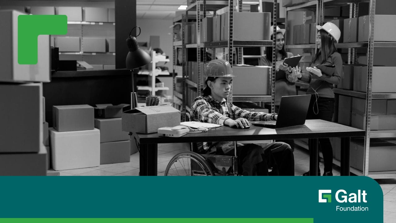 In a warehouse setting with shelves stocked with boxes, an individual in a wheelchair is sitting at a desk with a laptop in front of them, and two other people wearing hard hats are standing in the background.